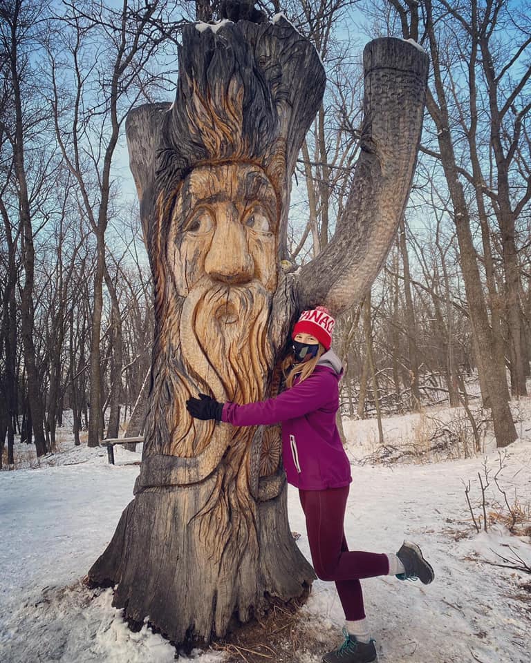 Image of a young women hugging a tall tree stump carved into the face of a wizard in winter.